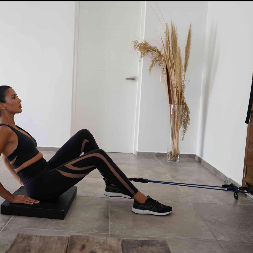 A woman in workout attire performing a Leg Curl Substitution with A90 Ankle Straps and a resistance band in a modern, minimalist room.