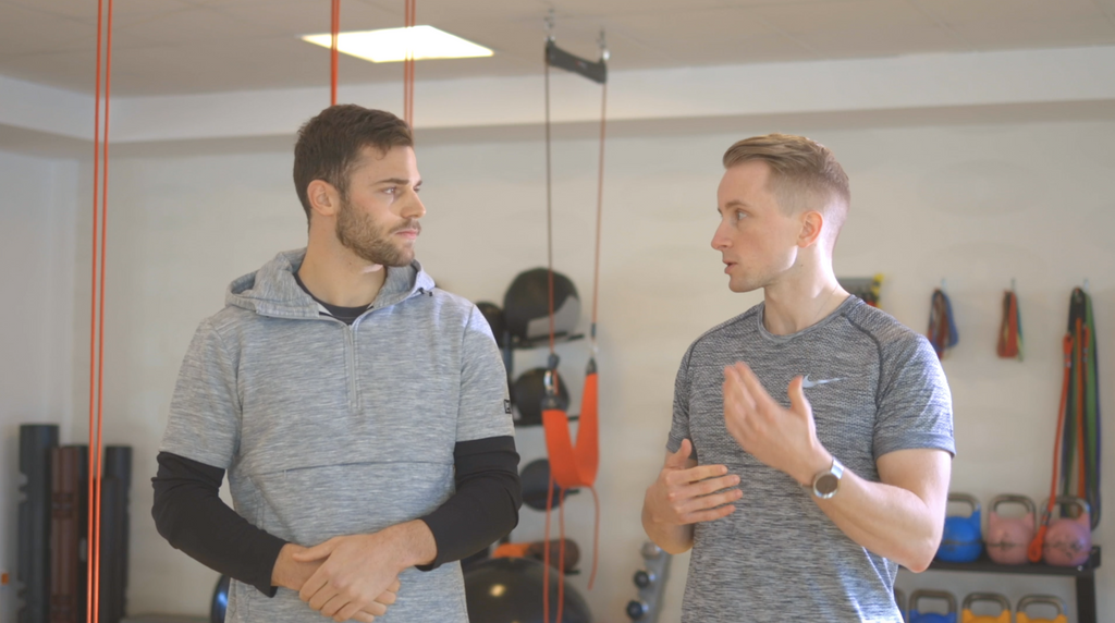 Two men having a discussion about injury prevention in a gym with A90 Prehab: Self-Screening & Corrective Exercises (Video Course) in the background.